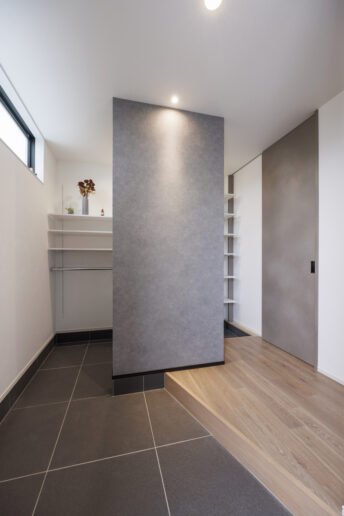 グレーを基調にコーディネートした玄関ホール。斜めに切った框と、天井まで届くハイドアが空間を広く見せます。シューズクローゼットの手前に設けた壁には、目隠しと空間のアクセントという2つの要素を持たせています。壁を境に、日常の動線と来客時の動線がシンプルに分られています。